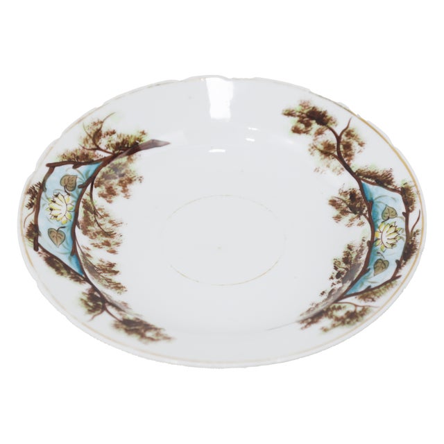 A delicate antique porcelain saucer featuring a hand-painted Asian landscape scene with lush trees, lotus flowers, and...
