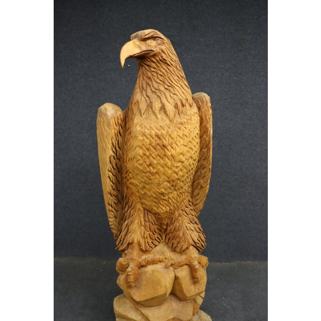 A large hand-carved pine sculpture depicting a bald eagle perched atop a stylized, saw-cut rock formation. Executed from a...