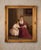 Vintage oil painting depicting a tender interior scene of a mother reading to a young child. The composition has a warm,...