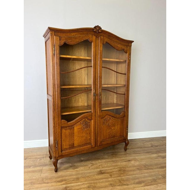 An elegant Vintage French Louis bookcase in oak, featuring glazed double doors, a shaped top, carved decorative details...
