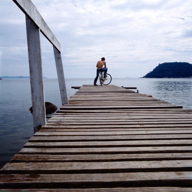 The image shows a cyclist on a wooden pier extending over the calm waters of Guanabara Bay. The cyclist, shirtless and...