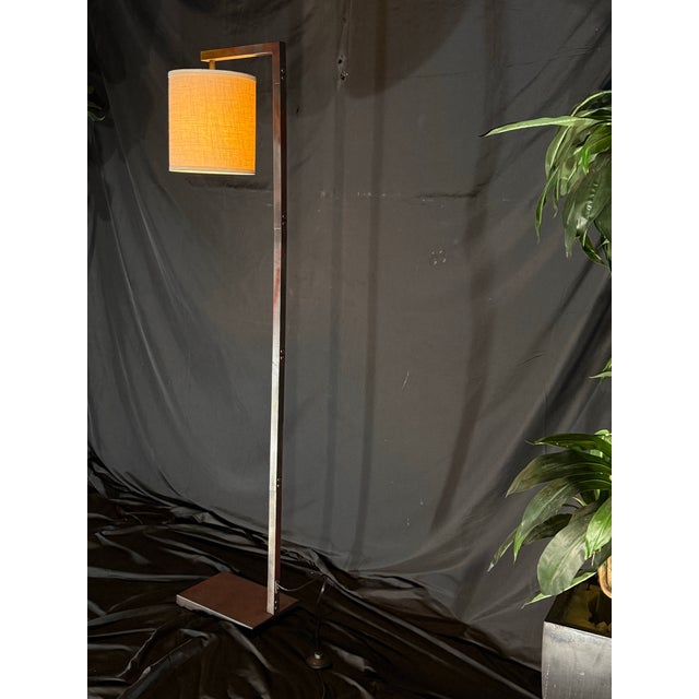 A modern minimalist floor lamp with a clean rectangular frame, square base, and cream fabric drum shade. The frame...