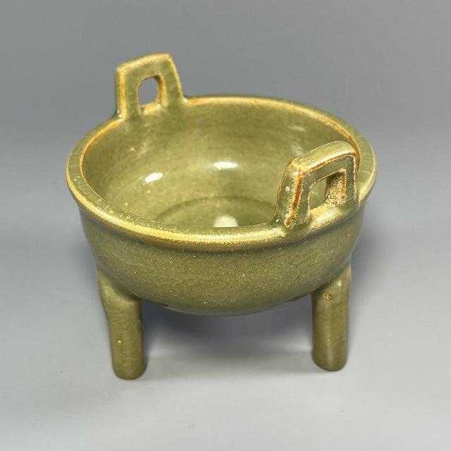 A vintage‑style Chinese Longquan celadon tripod censer, supported by three sturdy legs and fitted with two upright side...