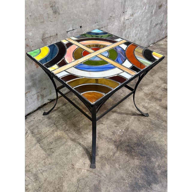 A striking vintage side table featuring a colorful stained glass top set within a sculptural wrought iron base. The...