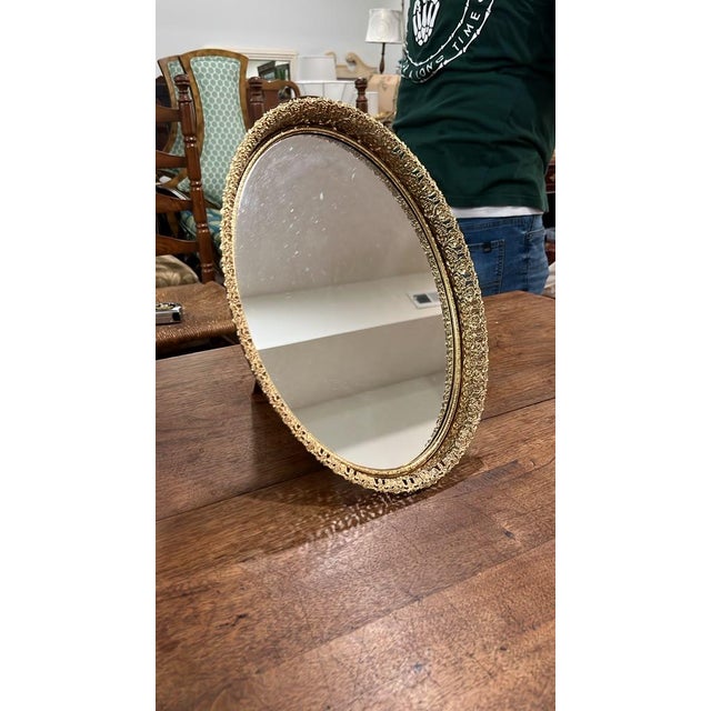 An ornate vintage oval tabletop vanity mirror featuring a detailed, pierced filigree gilt-metal rim. Designed with an...