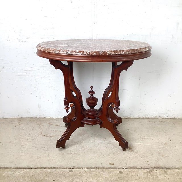 An elegant Victorian Revival center table featuring a richly veined oval marble top above an elaborate carved walnut-toned...