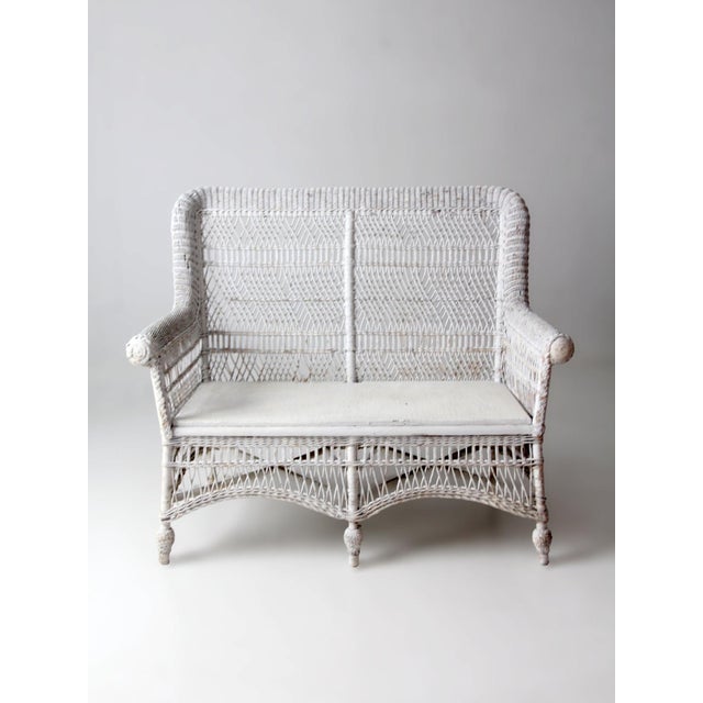 An early 20th-century wicker loveseat with a high woven back, gracefully rolled arms, and intricate open latticework. The...