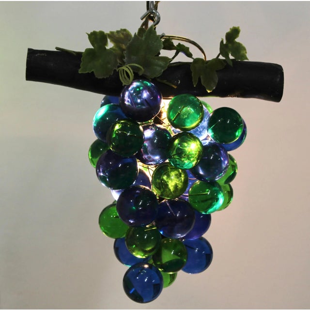 A bunch of large blue and green solid lucite grapes hanging from a brown stem with several small light bulbs within the...