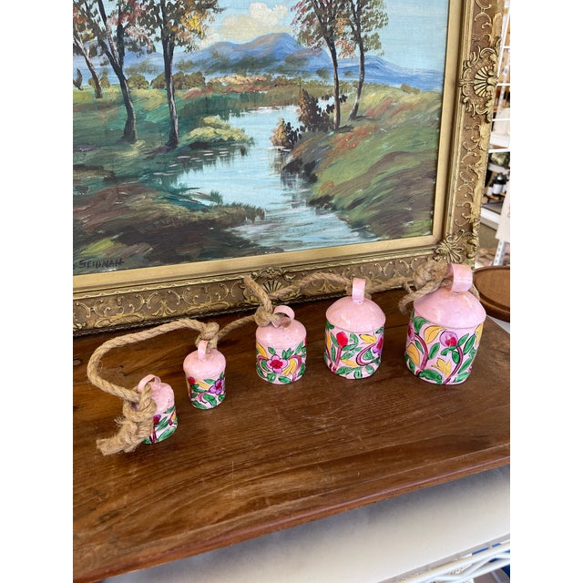 This vibrant decorative garland features a set of six hand-painted metal bells in graduated sizes, each adorned with a...