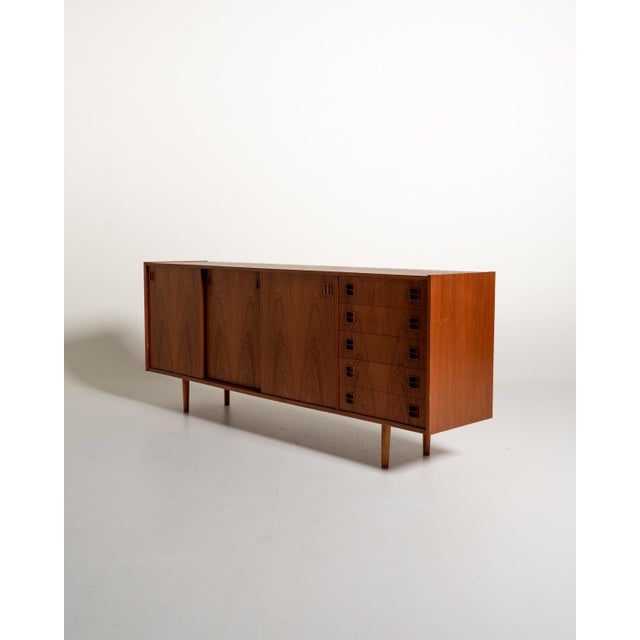 Danish-made teak sideboard from the 1960s, featuring three sliding doors and five side drawers. The front is accented by...