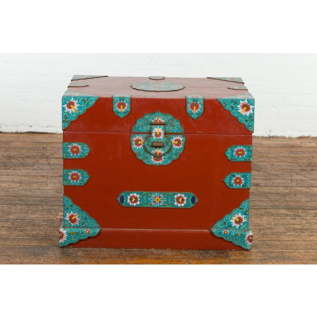 A vintage Chinese blanket chest from the mid 20th century with red lacquer ground, cloisonné décor and lateral handles....