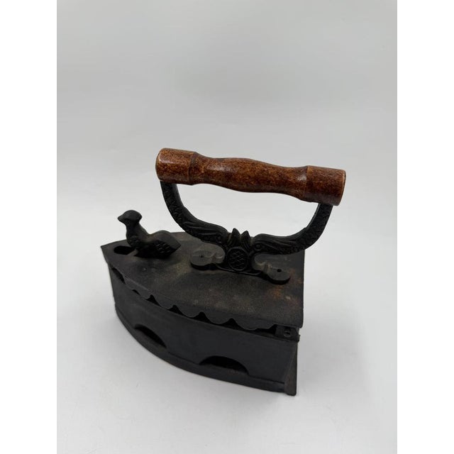Antique cast iron with a wooden handle and a decorative rooster figurine on the front. The surface is dark and shows age-...