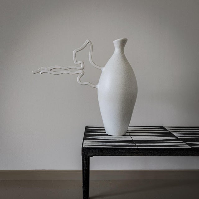White glazed ceramic vase with a smooth conical body and a long free-form handle on the side. The surface shows a fine...
