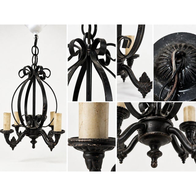 This chandelier features a sculptural black wrought-iron frame with elegant scrollwork, candle-style holders, and a...
