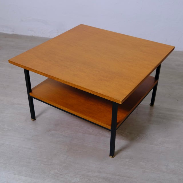 This vintage coffee table from the 1960s features a square top and a lower shelf in warm wood on a black metal frame....