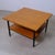 This vintage coffee table from the 1960s features a square top and a lower shelf in warm wood on a black metal frame....