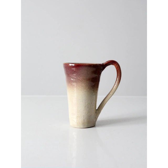 This vintage studio pottery mug is a weighty, hand-crafted ceramic piece with a creamy white glaze accented by a rich...