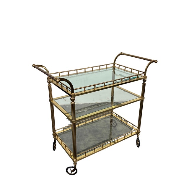 Elegant vintage Regency-style three-tier bar cart featuring a brass frame with clear glass shelves and oversized spoked...