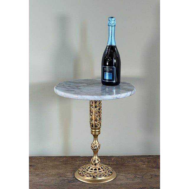 This vintage pedestal side table features a round white marble top with subtle gray veining set on an ornate cast brass...