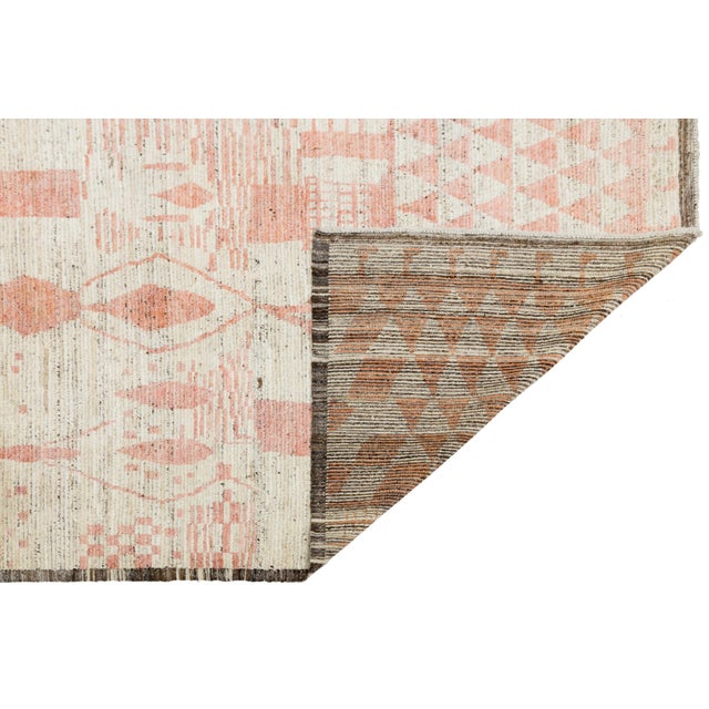 This modern Moroccan-style wool rug showcases a playful mix of abstract geometric motifs in soft pink and red tones over a...