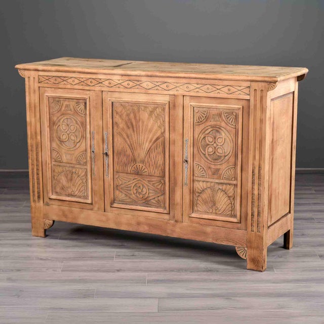 A three-door oak sideboard in natural, bare-wood finish, carved with Art Deco motifs. A central sunburst and wheat sheaf...
