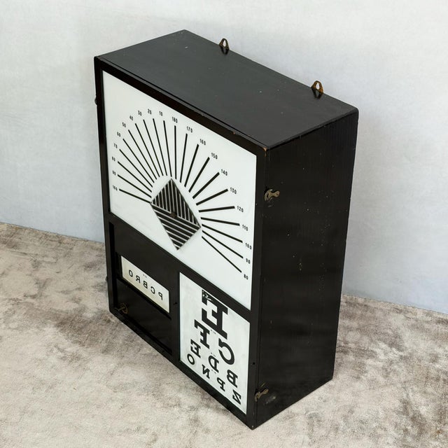A wall-mounted optician's test cabinet in a black-painted wooden case. Behind a hinged front frame there are three...
