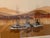 A serene sepia-toned lithograph of fishing trawlers at rest in a misty harbor, layered mountains fading behind them....