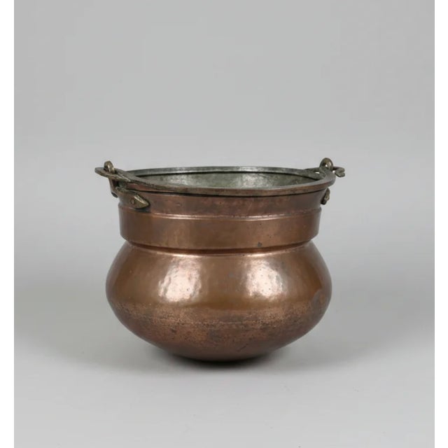 A substantial 19th-century hand-hammered copper cauldron of low, bulbous form, with a wide rolled rim and opposing iron...