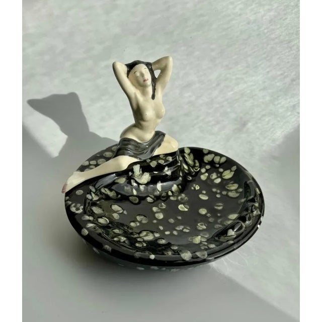 A striking mid-century ceramic ashtray featuring a sculptural nude female figure seated gracefully on the edge of a...
