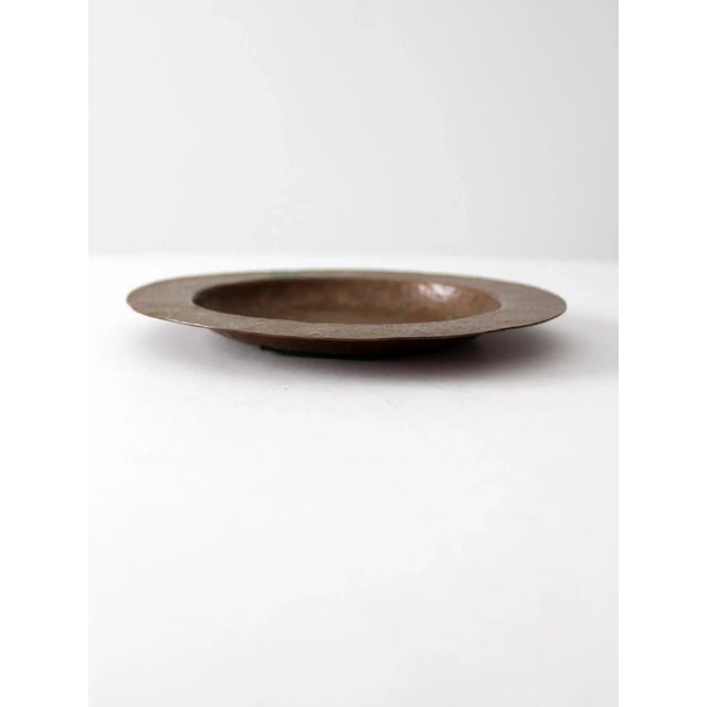 An antique hammered copper bowl showcasing a richly textured surface and a warm, aged patina. The shallow bowl features a...