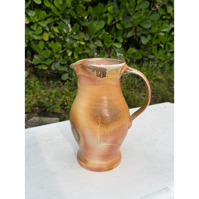 Vintage handcrafted studio pottery pitcher featuring a simple, organic form with a gently flared rim and elongated handle....