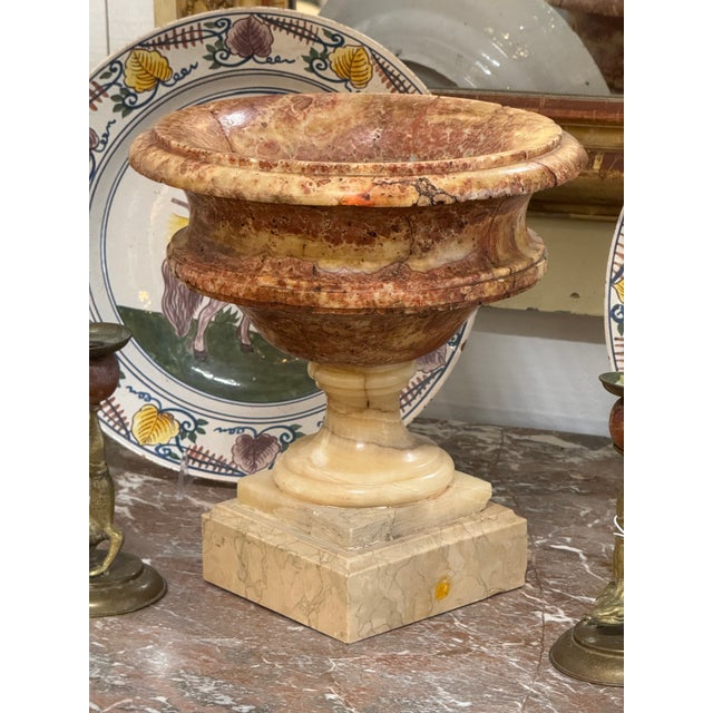 Marble urn with a flared rim and rounded body raised on a turned stem over a stepped square base. The stone shows varied...