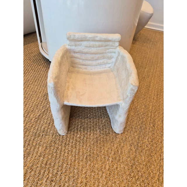 A ceramic sculpture of a chair. Made of clay and is glazed in a natural white. The finish has some texture which gives it...