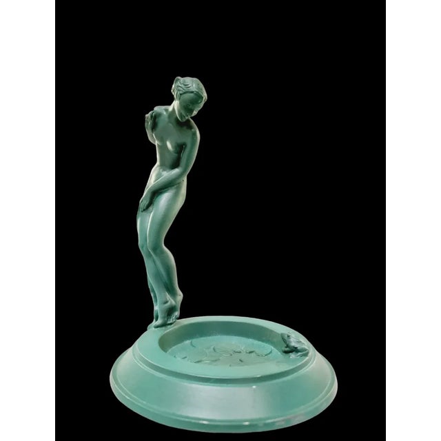 A nude woman is fearfully stepping into lake with water lilies and a cute frog. The figurine is covered with vibrant green...