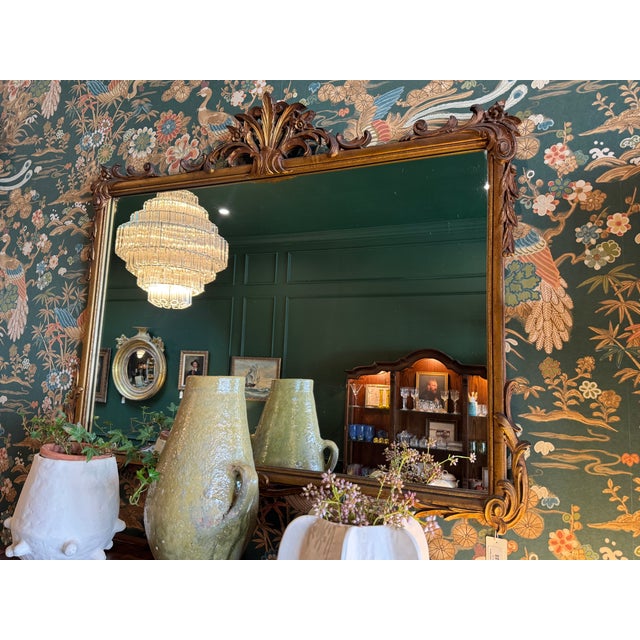Large, ornate rectangular mirror with a carved golden frame featuring an elaborate crest at the top center consisting of...