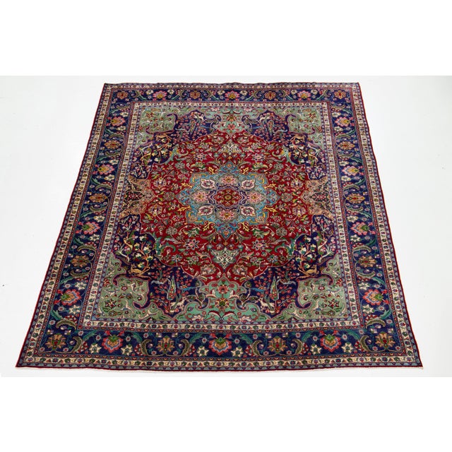 This handmade Tabriz red wool rug showcases a stunning rosette design, accented with vibrant hues of green, blue, and...