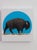 In "Blue Sunset," a solitary bison strides across a textured landscape, set against a striking blue circle that evokes the...