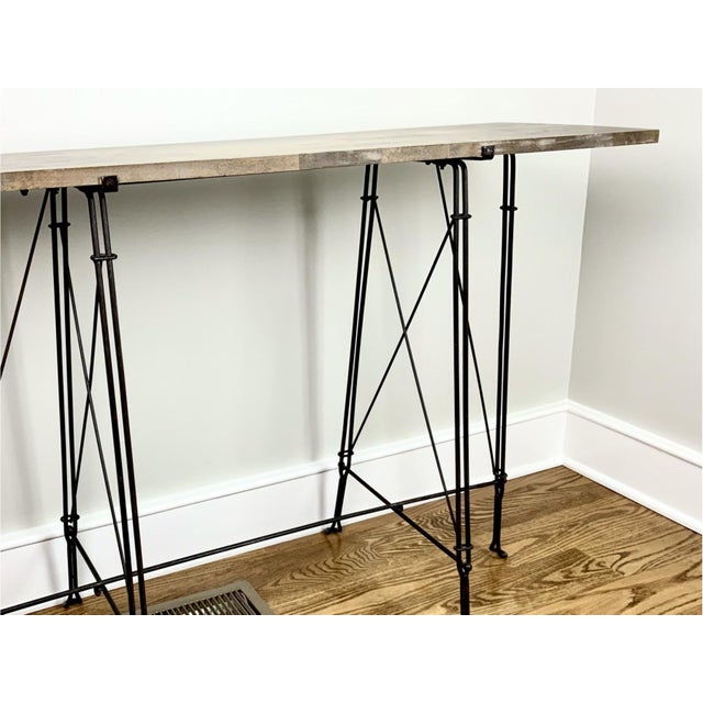A rectangular industrial style console table with a weathered, planked wooden top, ornate wrought iron base of slender,...