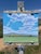 The painting depicts a serene landscape under expansive skies, where soft, wispy clouds drift lazily above rolling hills....