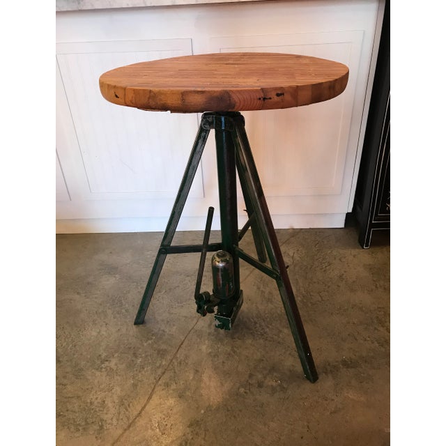Round industrial table with slatted wood top and steel tripod legs. It has a hydraulic mechanism at the bottom that adds a...