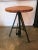 Round industrial table with slatted wood top and steel tripod legs. It has a hydraulic mechanism at the bottom that adds a...