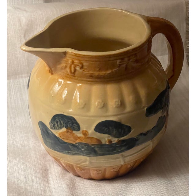 This Antique Roseville Woodlands Countryside Pitcher is an earthenware pottery piece from the 1910s, featuring a...