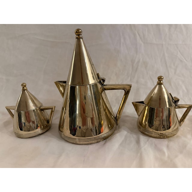 Art Deco silver-tone metal tea set comprising a teapot, sugar pot, and milk pot. The set features streamlined forms and...