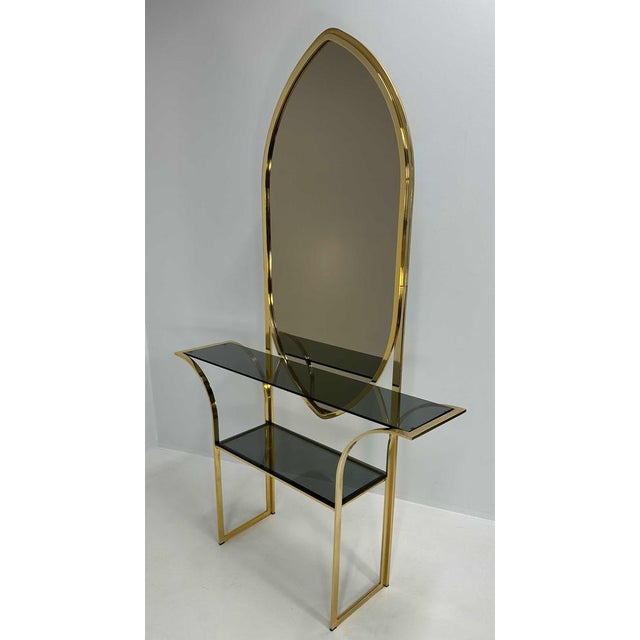 Italian console from the 1970s in brass with smoked glass shelves and a matching mirror. The structure features sinuous...
