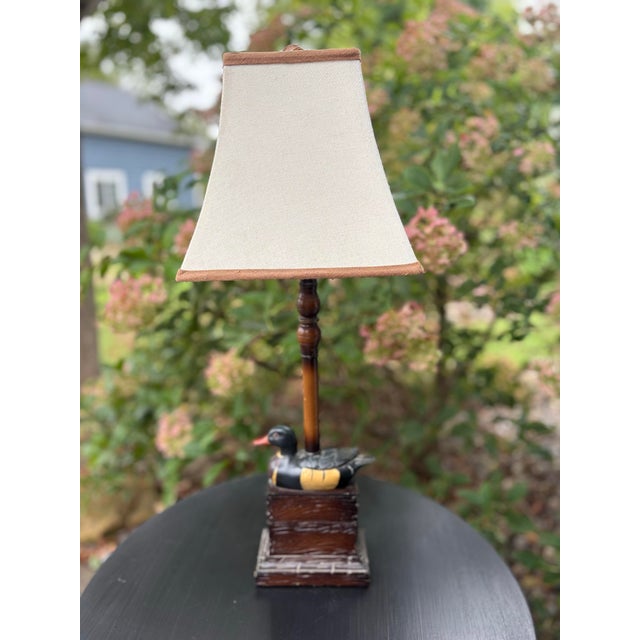 Charming vintage table lamp featuring a hand-painted duck perched at the base of a warm wood-tone column. The sculptural...