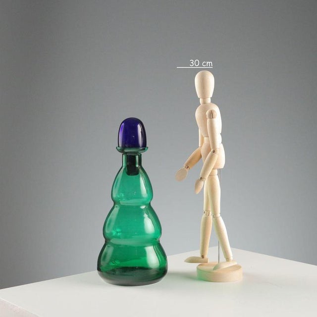 Green glass bottle with blue cap. Designer's signature. Bottle height 26 cm. Original box. This piece is attributed to the...