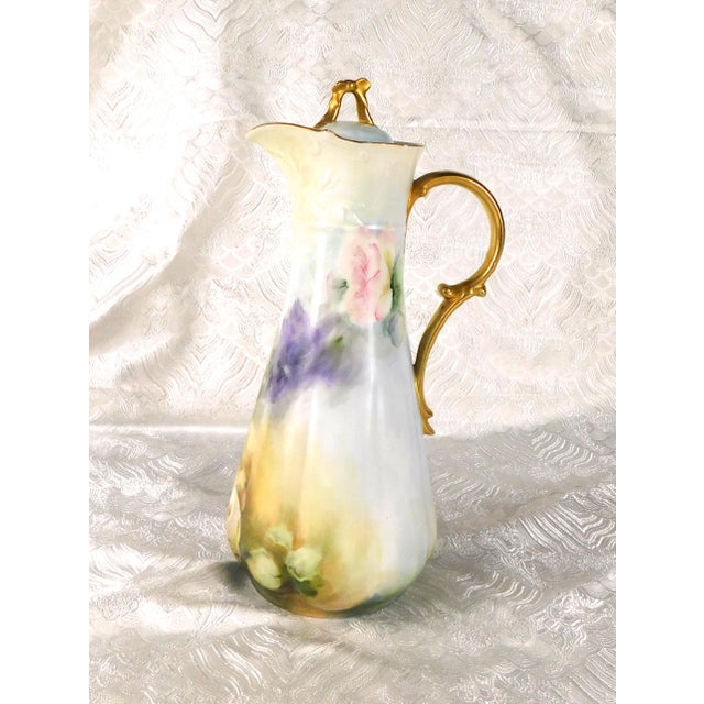 This is an unmarked but signed tall slender chocolate pot. The pot is white with a floral design on the left, but almost...
