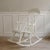 A white lacquered rocking chair in the style of Windsor, dating from the mid-20th century. The high back with its slender...