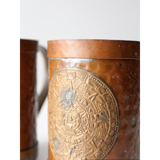A pair of vintage copper-plated mugs with a hammered surface and embossed Aztec calendar medallions. The warm copper tone...