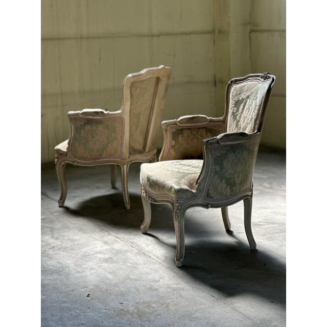 This is a pair of Louis XV style bergère chairs, elegant and stately in their form. The chairs feature a delicate, cream-...